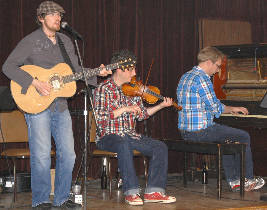 Photo of Jason MacDonald on vocals and guitar accompanied by Colin Grant on fiddle and Adam Young on piano